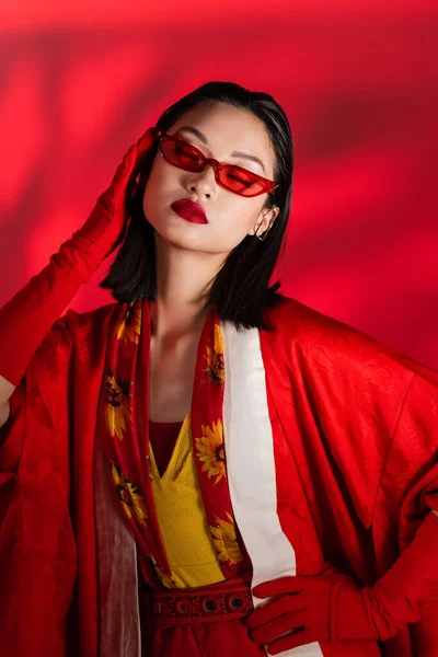 Moda asiática mulher no quimono capa e óculos de sol posando com olhos fechados no fundo vermelho com sombra — Fotografia de Stock