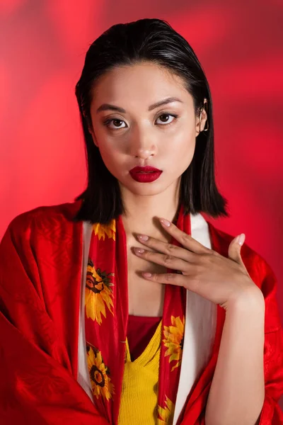 Morena mujer asiática en kimono capa y bufanda con decoración floral mirando a la cámara sobre fondo abstracto rojo — Stock Photo