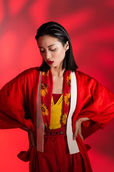 Brünette asiatische Frau in Halstuch mit floralem Dekor und Kimono-Umhang posiert mit den Händen an den Hüften auf abstraktem roten Hintergrund — Stockfoto