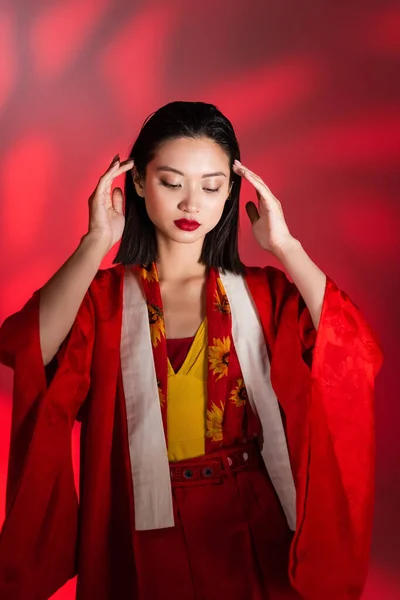 Elegante donna asiatica in mantello kimono rosso in posa con le mani vicino alla testa su sfondo astratto — Foto stock