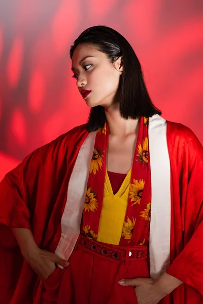 Morena mujer asiática en bufanda con estampado floral y capa de kimono posando con las manos en bolsillos sobre fondo con sombra roja - foto de stock
