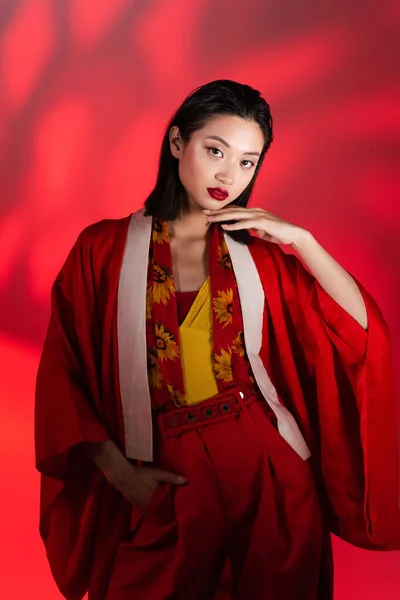Elegante mujer asiática en capa de kimono y bufanda con estampado floral de pie con mano en bolsillo sobre fondo rojo sombreado - foto de stock