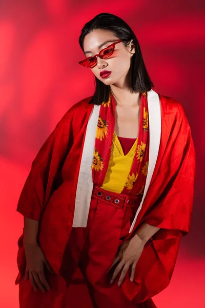 Модная азиатка в кимоно и солнцезащитных очках позирует на красном фоне с тенью — стоковое фото
