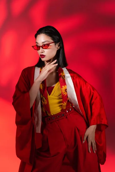 Jovem mulher asiática em óculos de sol e quimono capa posando no fundo abstrato com luz vermelha — Fotografia de Stock