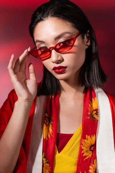 Retrato de morena mujer asiática tocando gafas de sol rojas y mirando a la cámara sobre fondo oscuro - foto de stock