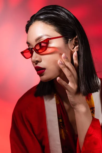 Азіатська жінка в модних сонцезахисних окулярах і вушному манжеті позує рукою біля обличчя на абстрактному фоні з червоним градієнтом — стокове фото