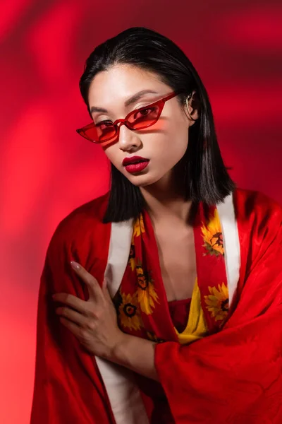 Молодая азиатка в кимоно и солнцезащитных очках смотрит в камеру на абстрактном красном фоне — стоковое фото