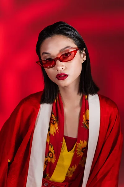 Morena mujer asiática en elegante kimono capa y gafas de sol con estilo mirando a la cámara sobre fondo rojo - foto de stock