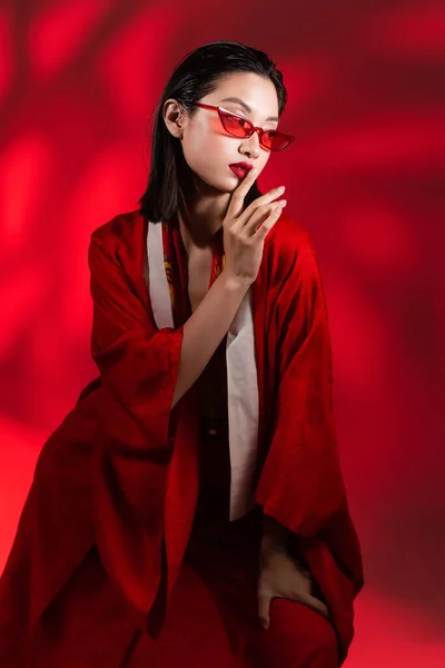 Morena mujer asiática en kimono capa y gafas de sol mostrando signo de silencio sobre fondo rojo con sombra - foto de stock