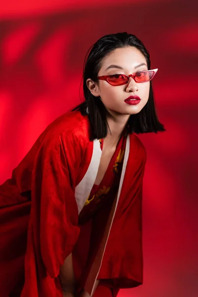 Brünette asiatische Frau mit stylischer Sonnenbrille und Kimono-Umhang schaut in die Kamera auf rotem Hintergrund — Stockfoto