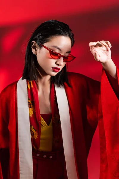 Elegante asiático mujer en elegante gafas de sol y kimono capa posando sobre rojo fondo con sombra - foto de stock