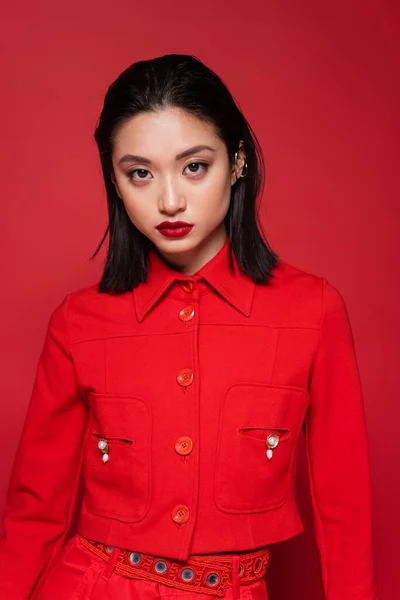 Morena mujer asiática con maquillaje usando elegante chaqueta y mirando a la cámara aislada en rojo - foto de stock