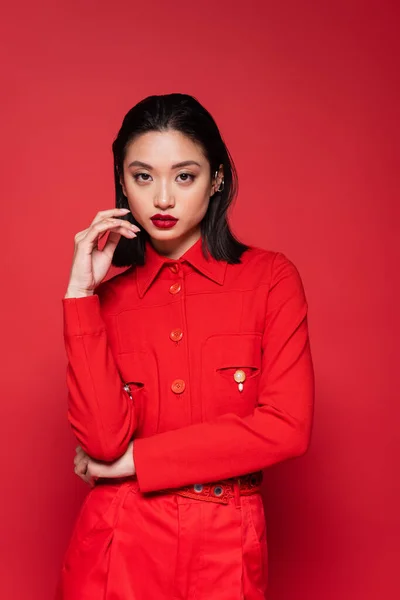 Bruna donna asiatica in giacca alla moda tenendo la mano vicino al viso isolato sul rosso — Foto stock