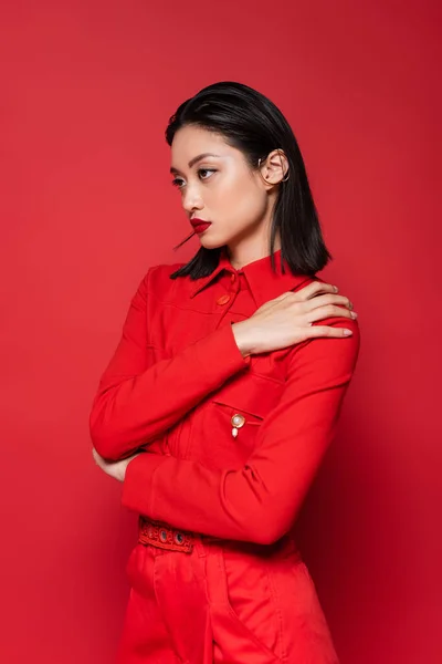 Alla moda donna asiatica in giacca posa con mano sulla spalla e guardando lontano isolato sul rosso — Foto stock