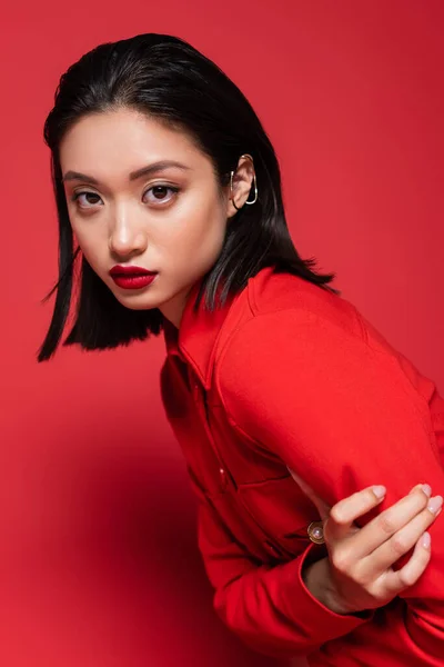 Retrato de morena mulher asiática com maquiagem e manguito de orelha olhando para a câmera no fundo vermelho — Fotografia de Stock