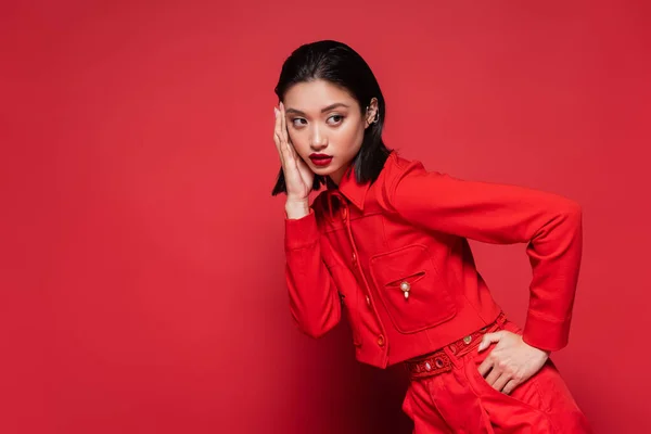 Jovem asiático modelo no elegante traje e orelha manguito tocando rosto e segurando mão no quadril no vermelho fundo — Fotografia de Stock