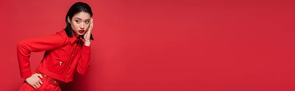 Elegante donna asiatica in elegante abbigliamento posa con mano sul fianco mentre tocca il viso su sfondo rosso, banner — Foto stock