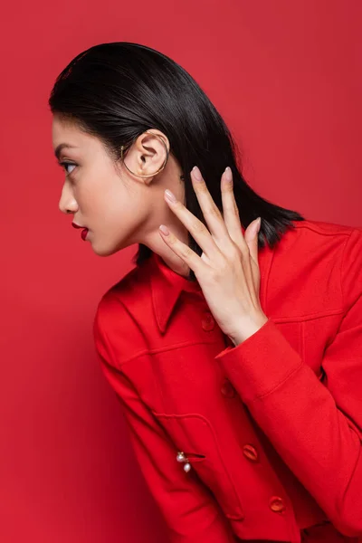 Elegante asiático mulher no casaco e orelha manguito segurando mão perto de pescoço e olhando para longe isolado no vermelho — Fotografia de Stock
