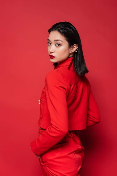 Femme asiatique dans des vêtements élégants debout avec la main dans la poche et en regardant la caméra sur fond rouge — Photo de stock
