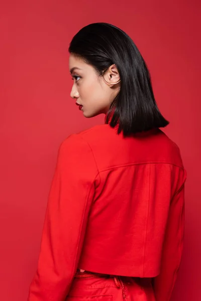 Joven asiático mujer en moda blazer y oreja manguito mirando lejos aislado en rojo - foto de stock