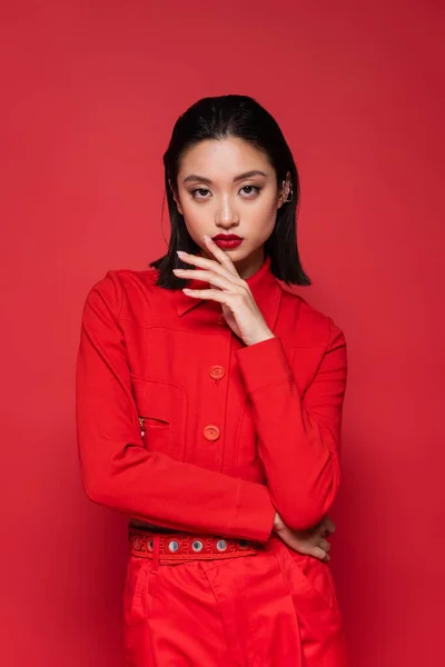 Брюнетка азіатська жінка в модному блістері тримає руку біля обличчя, дивлячись на камеру ізольовано на червоному — стокове фото