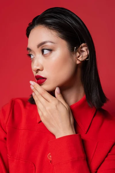 Porträt einer brünetten asiatischen Frau mit Ohrenmanschette und Make-up, die die Hand am Kinn hält, während sie isoliert auf Rot wegschaut — Stockfoto