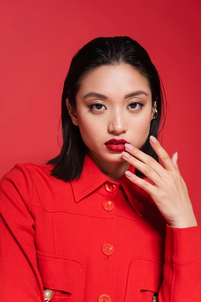 Retrato de bonita mujer asiática en elegante chaqueta mirando a la cámara y tocando los labios aislados en rojo - foto de stock