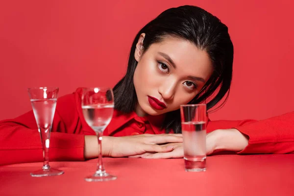 Portrait of brunette asian woman in jacket posing near different glasses with water isolated on red — Stock Photo