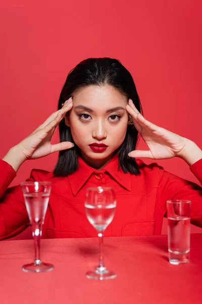 Portrait of asian woman in trendy jacket posing near glasses with clear water isolated on red — Stock Photo