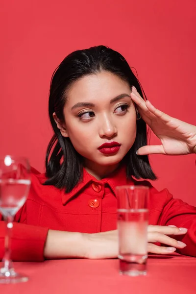 Stylish asian woman with makeup holding hand near face and looking away near blurred glasses with water isolated on red — Stock Photo