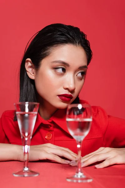 Retrato de morena mulher asiática com maquiagem olhando para longe perto de copos borrados de água isolada no vermelho — Fotografia de Stock