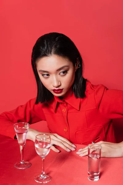 Morena mujer asiática en elegante chaqueta mirando lejos cerca de gafas con agua pura sobre fondo rojo - foto de stock