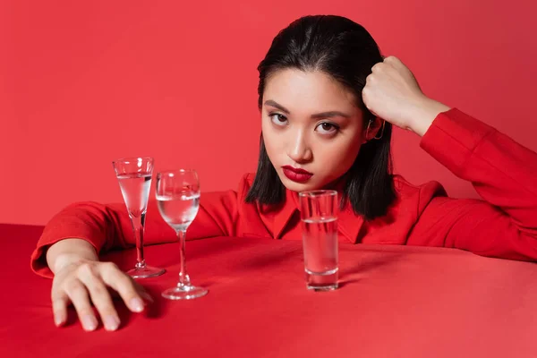 Young asian woman in jacket holding hand near head and looking at camera near glasses with water isolated on red — Stock Photo
