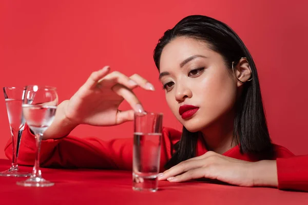 Jeune femme asiatique avec maquillage touchant verre avec de l'eau pure isolé sur rouge — Photo de stock