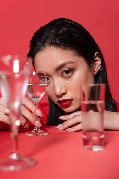 Portrait of asian woman with makeup and ear cuff near glasses of clear water on blurred foreground isolated on red — Stock Photo