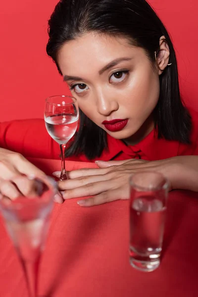 Brunette asian woman with makeup and ear cuff looking at camera near glasses with water on blurred foreground isolated on red — Stock Photo
