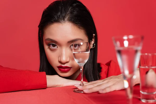 Portrait of young asian woman with makeup looking at camera near blurred glasses with water isolated on red — Stock Photo
