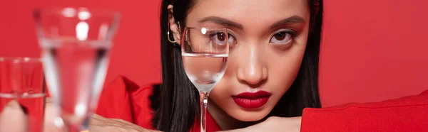 Портрет азиатки с брюнетками и макияжем возле размытых стаканов воды, изолированных на красном, баннер — стоковое фото