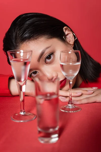 Portrait of brunette asian woman with ear cuff and makeup looking at camera near blurred glasses with water isolated on red — Stock Photo