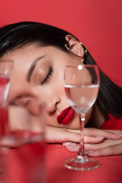Portrait of asian woman with makeup and closed eyes near glasses with pure water on blurred foreground isolated on red — Stock Photo