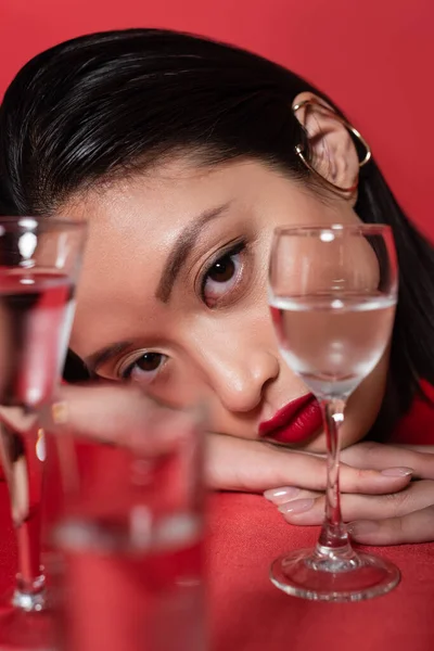 Ritratto di giovane modello asiatico con trucco guardando la fotocamera vicino vetri sfocati con acqua isolata su rosso — Foto stock