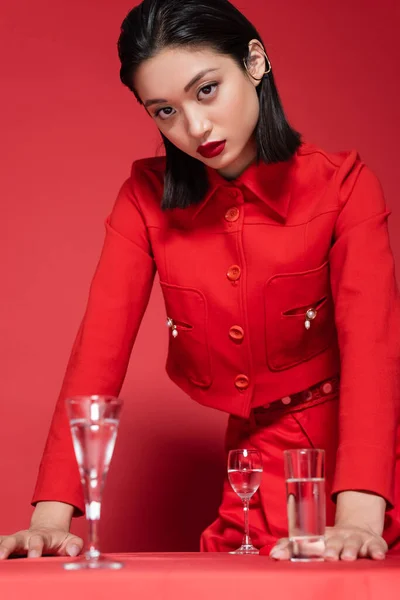 Morena mujer asiática en elegante chaqueta de pie cerca de gafas con agua limpia sobre fondo rojo - foto de stock