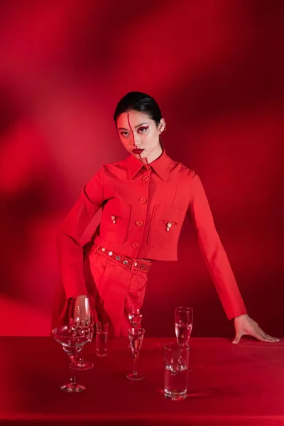 Trendige asiatische Frau mit artistischem Make-up, die in die Kamera schaut, nahe einer Brille mit klarem Wasser auf rotem Hintergrund mit Schatten — Stockfoto