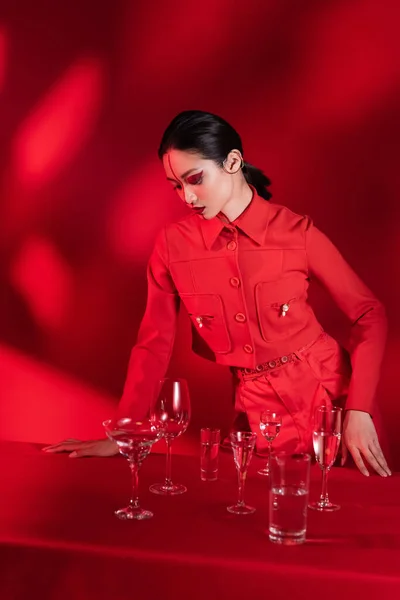 Asiatische Frau mit kreativem Make-up und Linie auf dem Gesicht posiert in stilvollem Anzug neben Brille mit Wasser auf rotem Hintergrund — Stockfoto
