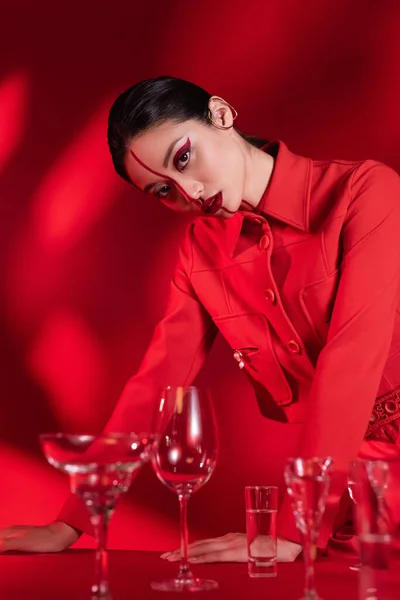 Modische asiatische Frau mit kreativem Make-up schaut in die Kamera in der Nähe von Gläsern mit Wasser auf rotem Hintergrund mit Schatten — Stockfoto