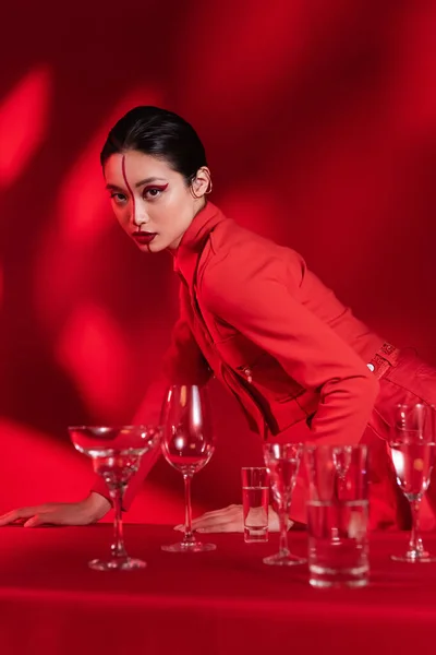 Модна азіатська жінка з художнім візитом на обличчі, розділена лінією, що позує біля різних окулярів на червоному тлі — стокове фото