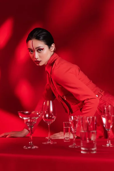 Trendy asiatische Frau mit künstlerischem Gesicht geteilt mit Linie posiert in der Nähe verschiedene Gläser auf rotem Hintergrund — Stockfoto