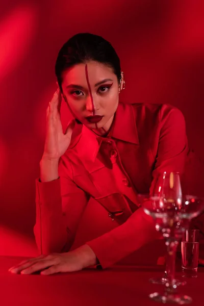 Femme asiatique avec visage créatif posant dans une veste à la mode près de verres flous avec de l'eau sur fond rouge ombragé — Photo de stock