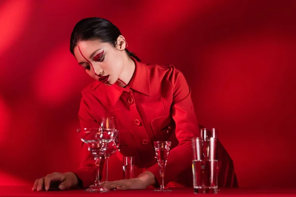 Femme asiatique avec maquillage artistique debout dans une veste à la mode près de différents verres avec de l'eau sur fond rouge — Photo de stock