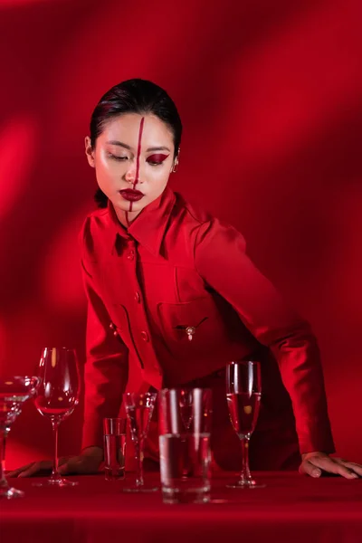 Asiatique femme avec lumineux maquillage sur visage divisé avec ligne posant près de verres d'eau sur fond rouge avec ombre — Photo de stock